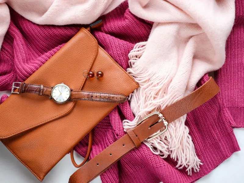 Bolsa, relógio, cinto e lenço dispostos sobre fundo rosa